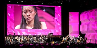 On the screen above the stage with the orchestra is the expressive face with closed eyes of a young pianist.
