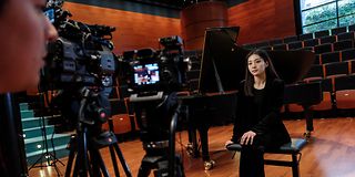 A black-haired woman in black clothes sits on a piano stool with her legs crossed, two cameras in front of her.