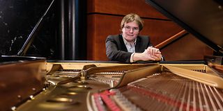 A young brown-haired man with glasses smiles through the open grand piano over the stringed golden soundboard.