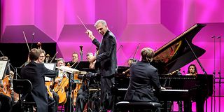 Conductor and pianist look at each other while making music with the orchestra. The walls of the stage are magenta.
