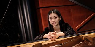 A black-haired woman smiles with her hands clasped together over the stringed soundboard of the grand piano.