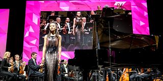 A pianist in a golden sequin dress stands at the edge of the stage to bow. Behind her sits the orchestra.