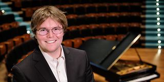 A young brown-haired man in glasses, a black suit and a white shirt laughs broadly into the camera. Behind him, the grand piano.