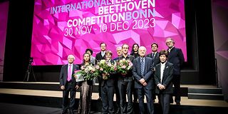 On the stage in front of the magenta screen of the competition are the jury and prize winners with bouquets of flowers.