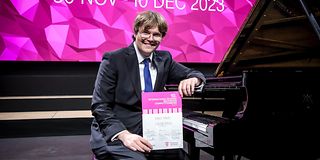 A young man in a black suit with a blue tie sits on the empty stage at the grand piano with a certificate in his hand and grins.
