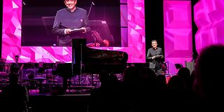 A man dressed in black gives a speech on stage in front of magenta walls.