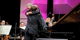 A young pianist and the conductor, both dressed in black, embrace on stage.