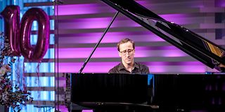 A young man with brown hair and glasses plays with his eyes closed on the grand piano in front of a magenta wall.