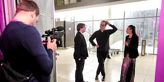 In a bright room with a glass wall, the three finalists stand carefree together, filmed by a cameraman.