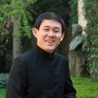 Ein asiatischer Mann mit schwarzem Hemd mit Kragen lächelt in einem Garten mit Beethovenbüste im Hintergrund in die Kamera.